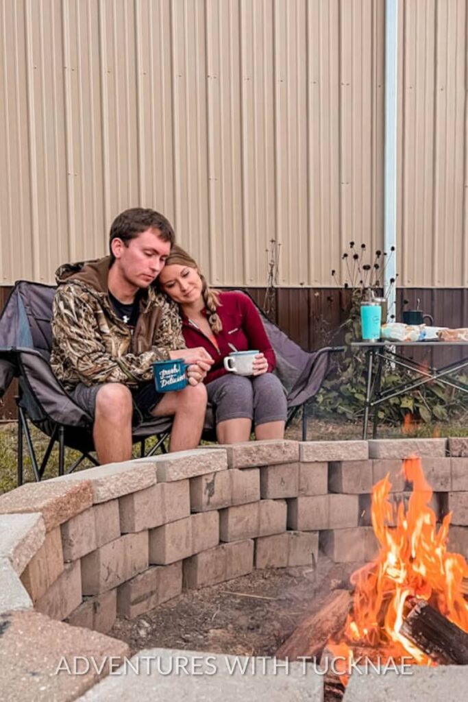 Tucker and Janae sit by a fire pit, bundled up and sipping from mugs while Janae rests her head on Tucker’s shoulder. A table with drinks and snacks is set up behind them beside a corrugated metal wall.