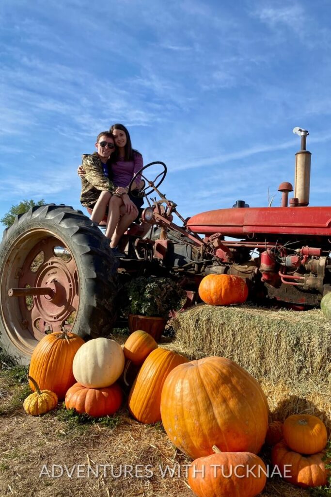 Tucker and Janae sit together on an old red tractor surrounded by hay bales and a variety of pumpkins in a festive autumn farm setting.