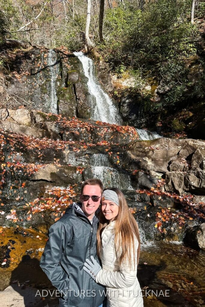 A couple poses in front of a waterfall cascading down mossy rocks covered in colorful autumn leaves in the Great Smoky Mountains. A romantic and nature-rich stop for fall RV destinations in the Southeast.
