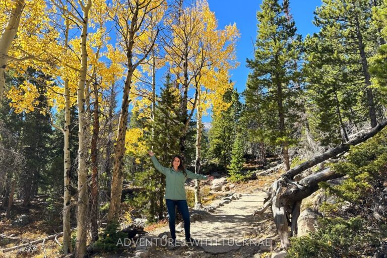Janae stands smiling with arms outstretched on a forested trail lined with golden aspens and evergreens in Rocky Mountain National Park. The crisp blue sky and vibrant fall colors capture the essence of scenic fall RV destinations in Colorado.
