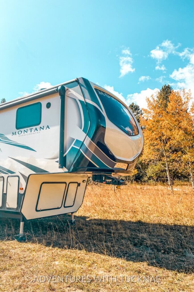 Close-up of a Montana fifth wheel RV parked on golden grass near Yellowstone National Park, with bright blue skies and orange-leaved trees in the background. A peaceful boondocking spot highlighting the natural beauty of fall RV destinations in the West.