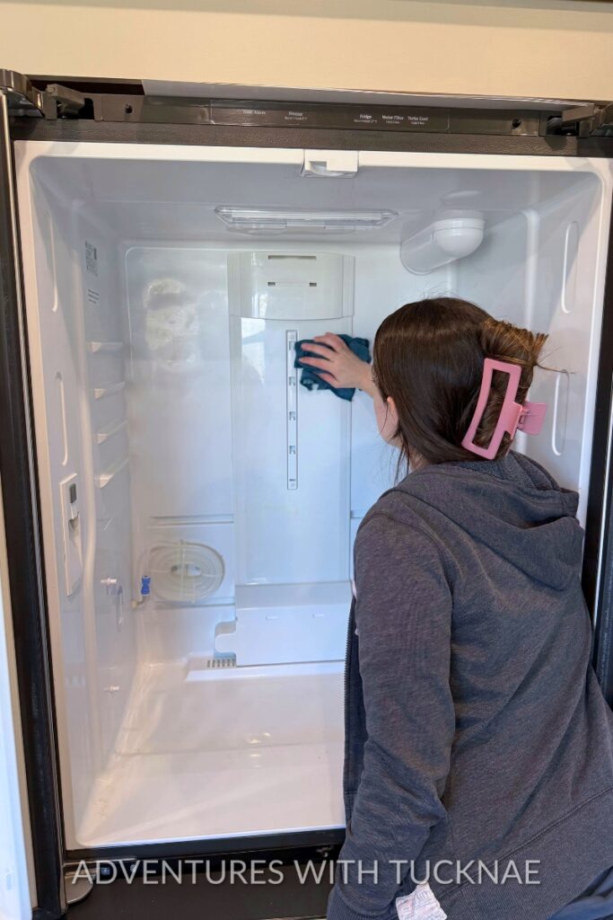 A woman cleaning inside an empty RV refrigerator with a microfiber cloth during RV spring cleaning. Open refrigerator doors and clean white interior show the appliance being sanitized and refreshed.