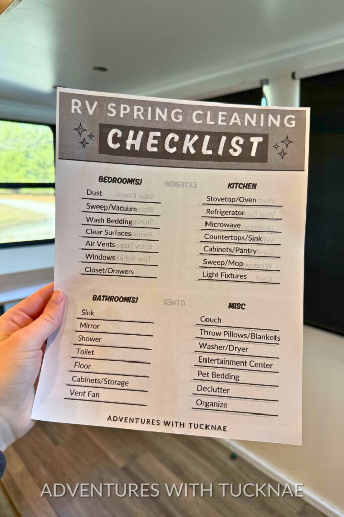 Hand holding a printable titled “RV Spring Cleaning Checklist” inside an RV living space. The checklist includes cleaning tasks for bedrooms, bathrooms, kitchen, and miscellaneous areas like washer and dryer, entertainment center, and pet bedding.