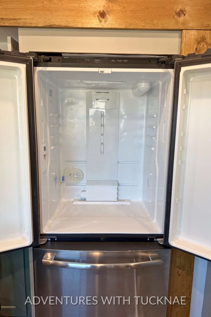Empty RV refrigerator with both doors open after being cleaned during RV spring cleaning. The bright white interior and cleared shelves show the fridge ready to be restocked for camping season.